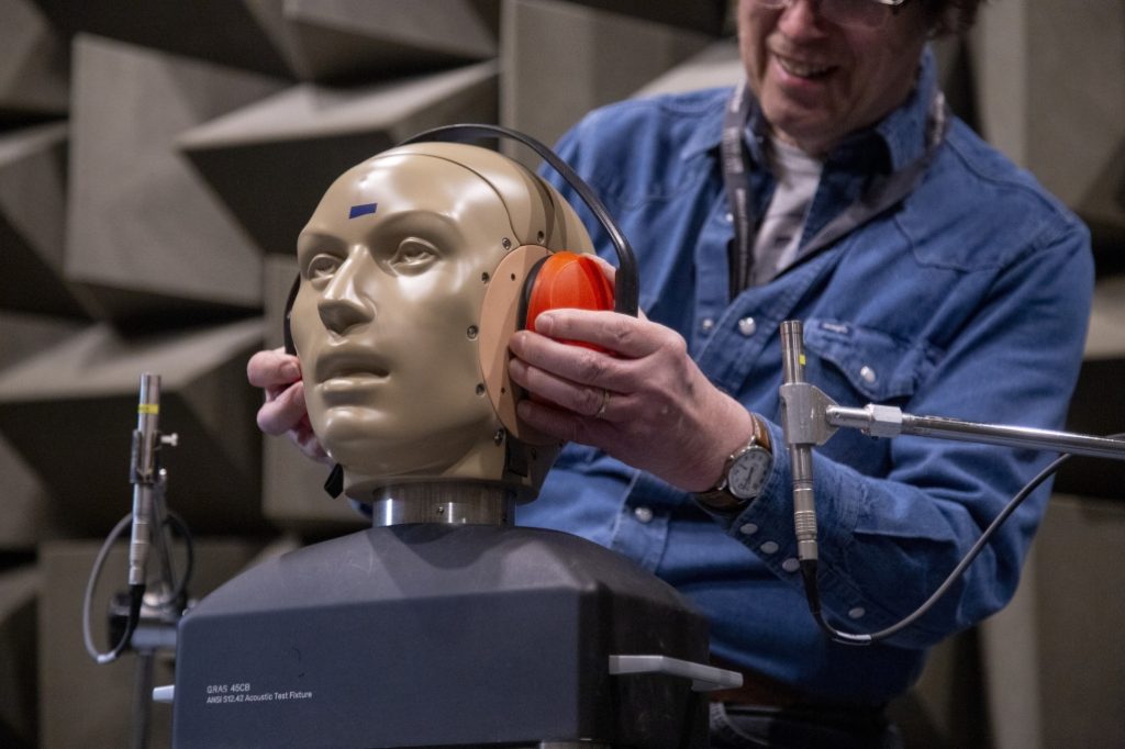 Person adjusting orange earmuffs on a life-sized dummy head used for acoustic testing in a sound-treated room. The mannequin head is mounted on equipment with attached microphones, and wedge-shaped acoustic foam panels line the walls in the background.