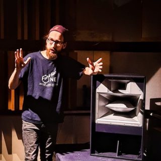 James Hipperson standing indoors next to a large loudspeaker cabinet, gesturing with both hands as if explaining or demonstrating something. The space features vertical wooden acoustic panels, and strong directional lighting casts shadows across the scene.