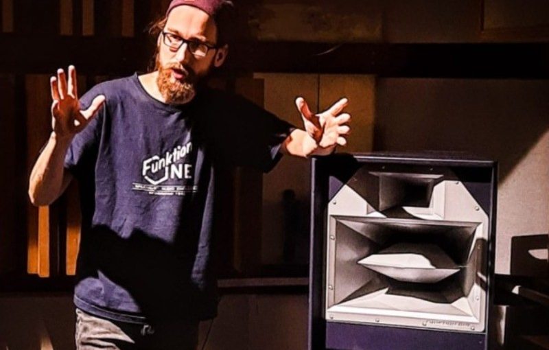 James Hipperson standing indoors next to a large loudspeaker cabinet, gesturing with both hands as if explaining or demonstrating something. The space features vertical wooden acoustic panels, and strong directional lighting casts shadows across the scene.