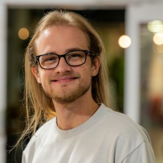 Image of Thomas Stolarski. Close-up portrait of a person with long light hair wearing dark-framed glasses and a light grey T-shirt, standing indoors and facing slightly toward the camera. The background is softly blurred with warm circular lights and glass panels, suggesting a relaxed indoor social or communal setting.