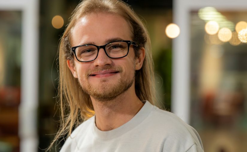 Image of Thomas Stolarski. Close-up portrait of a person with long light hair wearing dark-framed glasses and a light grey T-shirt, standing indoors and facing slightly toward the camera. The background is softly blurred with warm circular lights and glass panels, suggesting a relaxed indoor social or communal setting.