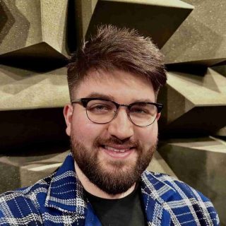 Close-up portrait of a person, Max Ellis, wearing glasses and a patterned jacket, standing in front of acoustic foam panels arranged in a geometric, wedge-shaped pattern typically used in sound testing environments.