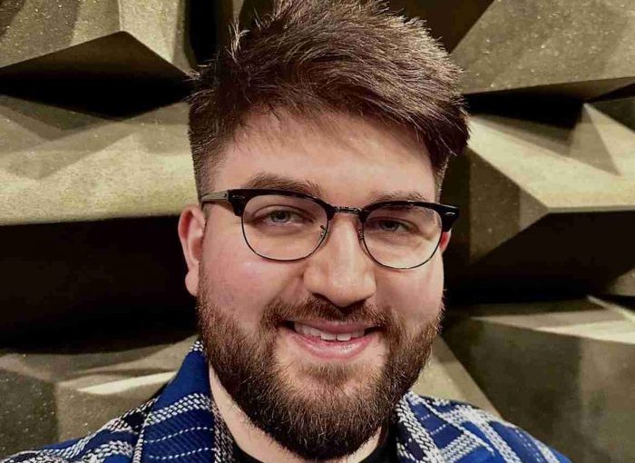 Close-up portrait of a person, Max Ellis, wearing glasses and a patterned jacket, standing in front of acoustic foam panels arranged in a geometric, wedge-shaped pattern typically used in sound testing environments.