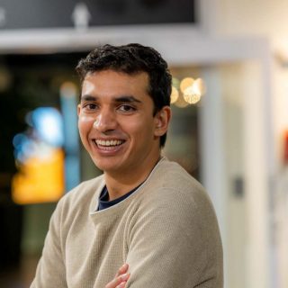 Image of Ahmed Khan. Close-up portrait of a person wearing a light beige textured sweater, turned slightly to one side with arms loosely crossed. The indoor background is softly blurred, featuring warm circular lights and glass panels, suggesting a relaxed social or communal setting.
