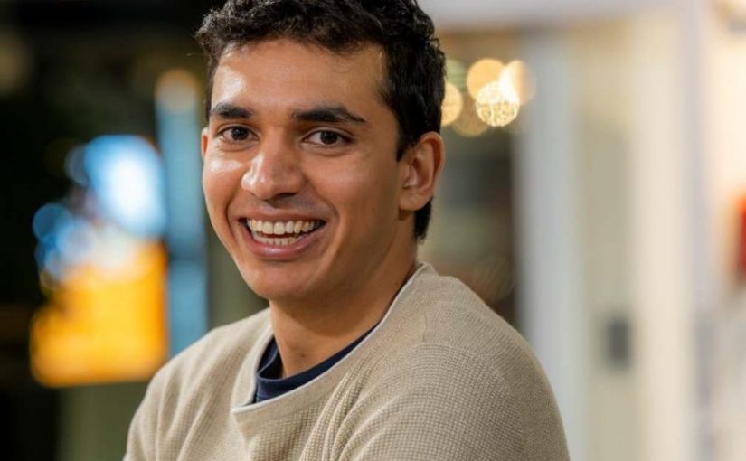 Image of Ahmed Khan. Close-up portrait of a person wearing a light beige textured sweater, turned slightly to one side with arms loosely crossed. The indoor background is softly blurred, featuring warm circular lights and glass panels, suggesting a relaxed social or communal setting.