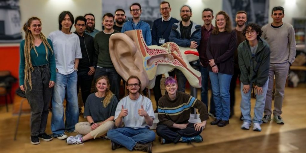 Group of around fifteen people, cohort 1, posed indoors around a large anatomical model of the human ear, which is centrally displayed and cross-sectioned to show internal structures. The group is arranged in two rows, with some standing behind the model and several seated on the floor in front. The room has a wooden floor and a softly blurred background with shelves and equipment, suggesting an academic or lab setting.