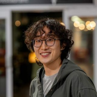 Image of Deokki Min. Close-up portrait of a person wearing round glasses and a casual jacket layered over a light top, photographed in an indoor setting. The background is softly blurred with warm, circular lights and glass panels, suggesting an informal social or communal space.