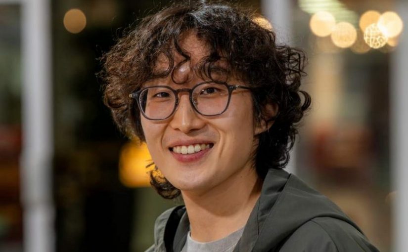 Image of Deokki Min. Close-up portrait of a person wearing round glasses and a casual jacket layered over a light top, photographed in an indoor setting. The background is softly blurred with warm, circular lights and glass panels, suggesting an informal social or communal space.