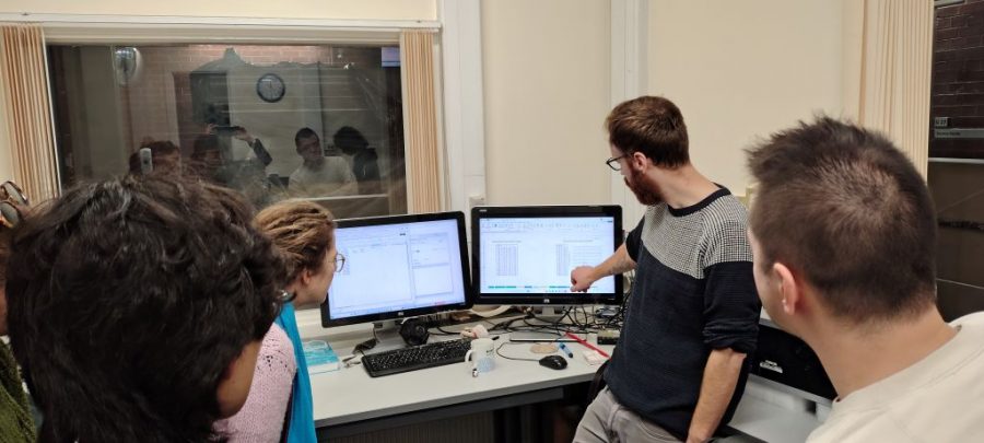 Small group gathered around a desk in a lab, with one person pointing at data displayed on dual computer monitors while others look on through a viewing window into a separate room.