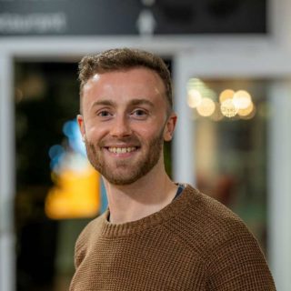 Image of Jack Hallybone. Close-up portrait of a person wearing a brown knitted sweater, facing slightly to one side. The indoor background is softly blurred, featuring warm, circular lights and glass panels, suggesting a relaxed social or communal environment.