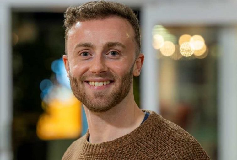 Image of Jack Hallybone. Close-up portrait of a person wearing a brown knitted sweater, facing slightly to one side. The indoor background is softly blurred, featuring warm, circular lights and glass panels, suggesting a relaxed social or communal environment.