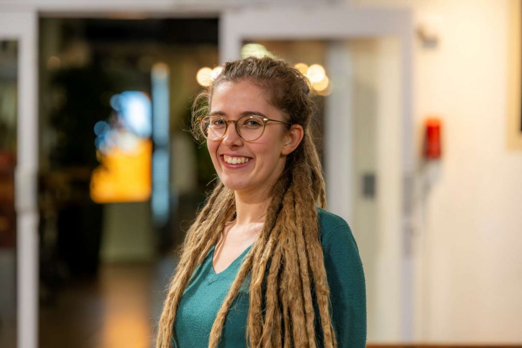 Image of Katie Salter. Close-up portrait of a person wearing glasses and a green top, with long dreadlocks draped over both shoulders. The person stands indoors in front of a softly blurred background with warm lighting and circular light reflections, suggesting an informal interior setting.