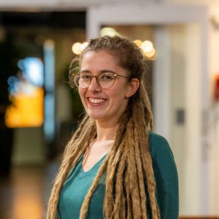 Image of Katie Salter. Close-up portrait of a person wearing glasses and a green top, with long dreadlocks draped over both shoulders. The person stands indoors in front of a softly blurred background with warm lighting and circular light reflections, suggesting an informal interior setting.