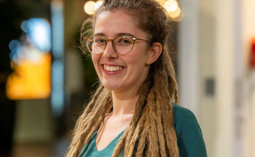 Image of Katie Salter. Close-up portrait of a person wearing glasses and a green top, with long dreadlocks draped over both shoulders. The person stands indoors in front of a softly blurred background with warm lighting and circular light reflections, suggesting an informal interior setting.