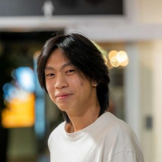 Image of Lucas Lee Zheng Xi. Close-up portrait of a person with medium-length dark hair wearing a plain white T-shirt, standing indoors and turned slightly toward the camera. The background is softly blurred with warm circular lights and glass panels, suggesting a relaxed indoor social or communal setting.