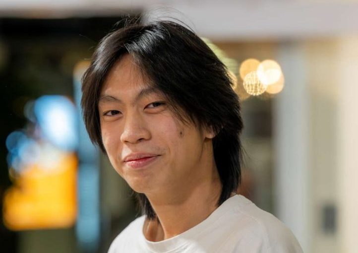 Image of Lucas Lee Zheng Xi. Close-up portrait of a person with medium-length dark hair wearing a plain white T-shirt, standing indoors and turned slightly toward the camera. The background is softly blurred with warm circular lights and glass panels, suggesting a relaxed indoor social or communal setting.