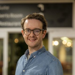 Image of Matt Gray. Close-up portrait of a person wearing dark-framed glasses and a light blue denim button-up shirt, positioned slightly angled toward the camera. The image is taken indoors, with a softly blurred background featuring warm circular lights, glass panels, and a dark overhead sign, suggesting a relaxed social or communal environment.
