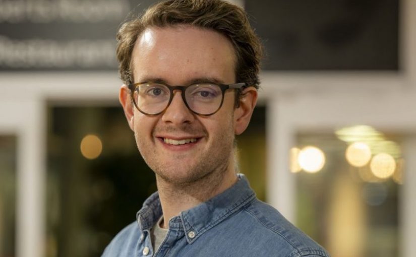 Image of Matt Gray. Close-up portrait of a person wearing dark-framed glasses and a light blue denim button-up shirt, positioned slightly angled toward the camera. The image is taken indoors, with a softly blurred background featuring warm circular lights, glass panels, and a dark overhead sign, suggesting a relaxed social or communal environment.