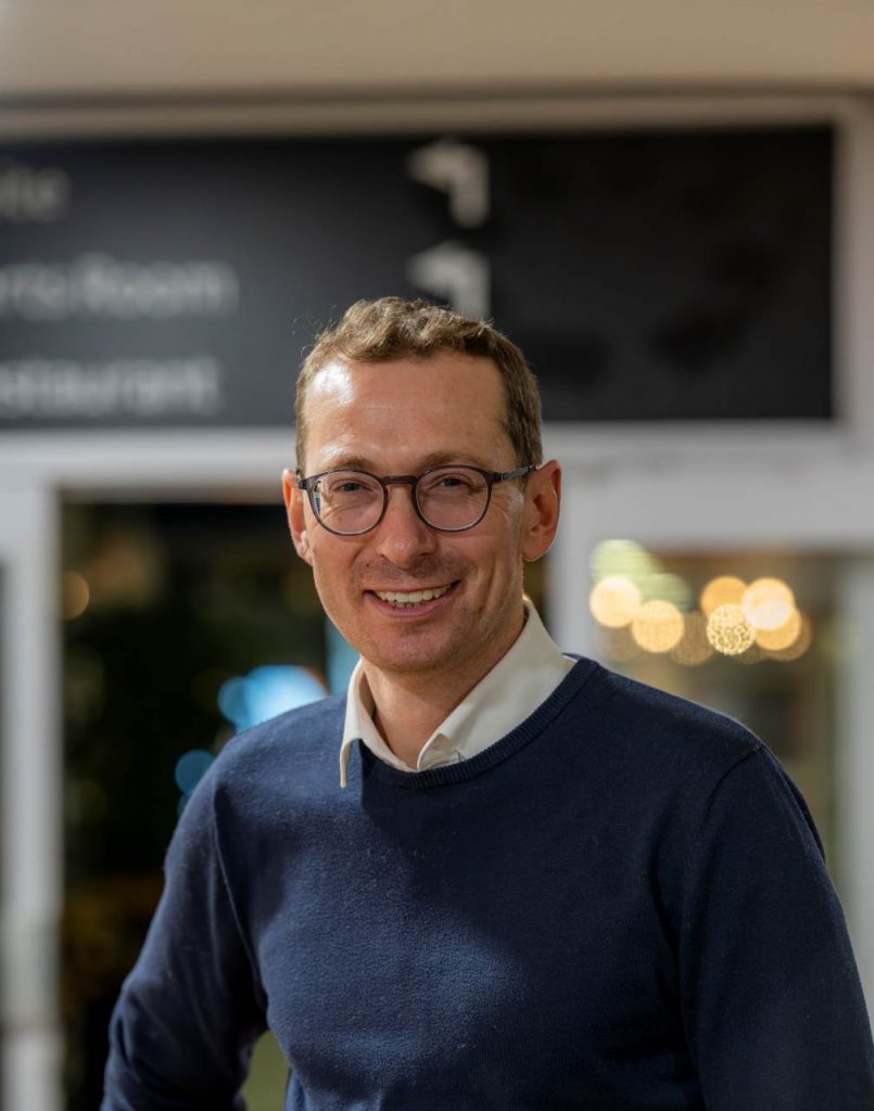 Image of Matt Torjussen. Close-up portrait of a person wearing round glasses and a navy jumper over a light collared shirt, standing indoors and facing slightly toward the camera. The background is softly blurred with warm circular lights and glass panels, suggesting a relaxed indoor setting.