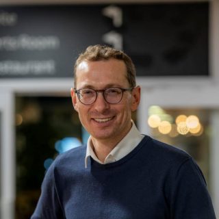 Image of Matt Torjussen. Close-up portrait of a person wearing round glasses and a navy jumper over a light collared shirt, standing indoors and facing slightly toward the camera. The background is softly blurred with warm circular lights and glass panels, suggesting a relaxed indoor setting.