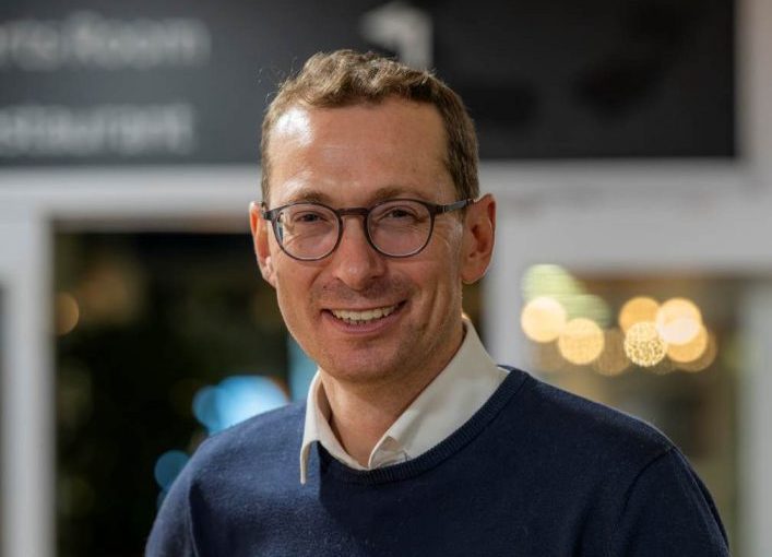 Image of Matt Torjussen. Close-up portrait of a person wearing round glasses and a navy jumper over a light collared shirt, standing indoors and facing slightly toward the camera. The background is softly blurred with warm circular lights and glass panels, suggesting a relaxed indoor setting.