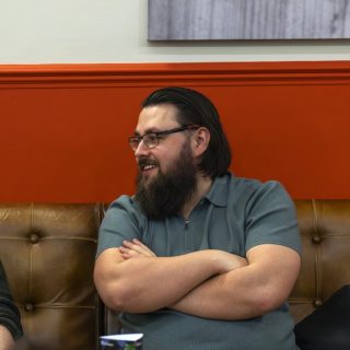 Image of Oscar Carter described as a person with glasses and a beard sitting on a brown leather bench with arms crossed, turned to the side in conversation. The background features a bold orange wall with a framed picture above, suggesting a casual indoor social or meeting space.