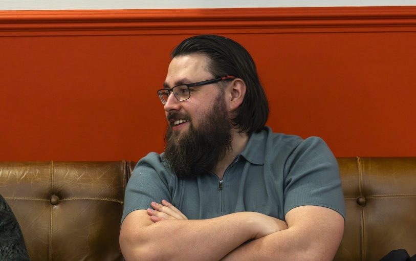 Image of Oscar Carter described as a person with glasses and a beard sitting on a brown leather bench with arms crossed, turned to the side in conversation. The background features a bold orange wall with a framed picture above, suggesting a casual indoor social or meeting space.