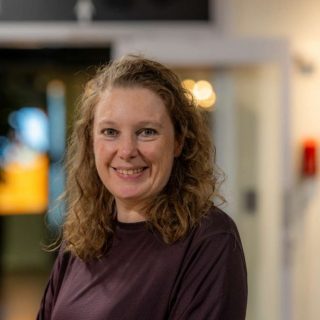 Image of Rebecca Hutt. Close-up portrait of a person with shoulder-length curly hair wearing a dark maroon long-sleeve top, positioned slightly angled toward the camera. The indoor background is softly blurred, showing warm circular lights and glass panels, suggesting a relaxed social or communal setting.