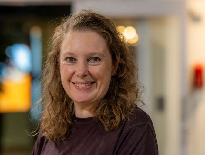 Image of Rebecca Hutt. Close-up portrait of a person with shoulder-length curly hair wearing a dark maroon long-sleeve top, positioned slightly angled toward the camera. The indoor background is softly blurred, showing warm circular lights and glass panels, suggesting a relaxed social or communal setting.