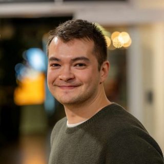 Image of Sean Cragg. Close-up portrait of a person wearing a dark green textured sweater, turned slightly to the side. The indoor background is softly blurred with warm circular lights and glass panels, suggesting a relaxed social or communal setting.