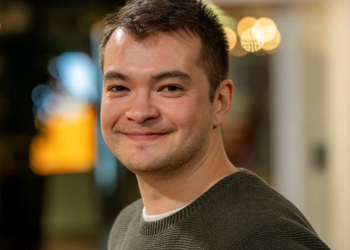 Image of Sean Cragg. Close-up portrait of a person wearing a dark green textured sweater, turned slightly to the side. The indoor background is softly blurred with warm circular lights and glass panels, suggesting a relaxed social or communal setting.