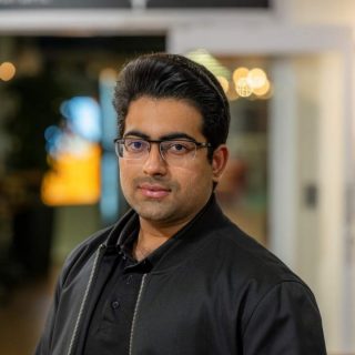 Image of Shahmir Panjwani. Close-up portrait of a person wearing rectangular glasses and a black jacket over a dark shirt, facing slightly toward the camera. The image is taken indoors, with a softly blurred background featuring warm, circular lights and glass panels, suggesting a relaxed social or communal environment.