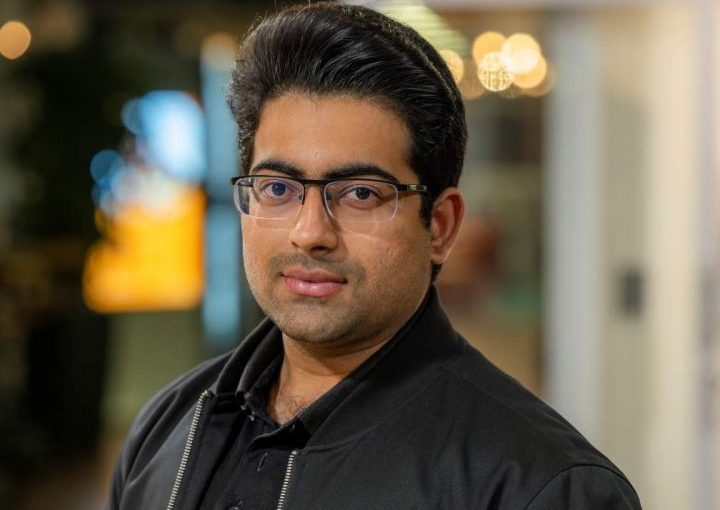 Image of Shahmir Panjwani. Close-up portrait of a person wearing rectangular glasses and a black jacket over a dark shirt, facing slightly toward the camera. The image is taken indoors, with a softly blurred background featuring warm, circular lights and glass panels, suggesting a relaxed social or communal environment.