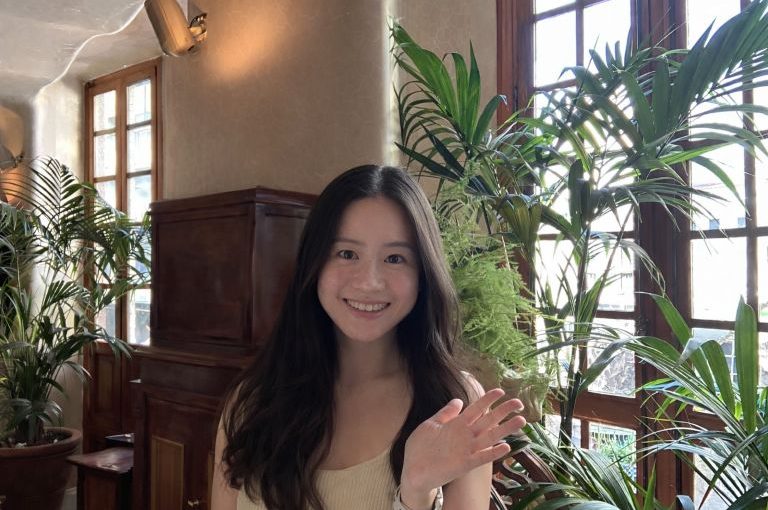 Image of Shumin Que. Close-up portrait of a person with long dark hair, raising one hand in a wave while standing indoors. The setting includes large green houseplants, wooden furniture, and a window allowing natural light, creating a bright and relaxed atmosphere.