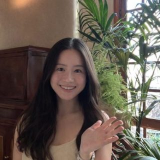 Image of Shumin Que. Close-up portrait of a person with long dark hair, raising one hand in a wave while standing indoors. The setting includes large green houseplants, wooden furniture, and a window allowing natural light, creating a bright and relaxed atmosphere.