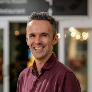 Image of Stuart Cumming. Close-up portrait of a person wearing a burgundy long-sleeve shirt, standing indoors and turned slightly toward the camera. The background is softly blurred with warm, circular lights and glass panels, suggesting a relaxed indoor social or communal environment.