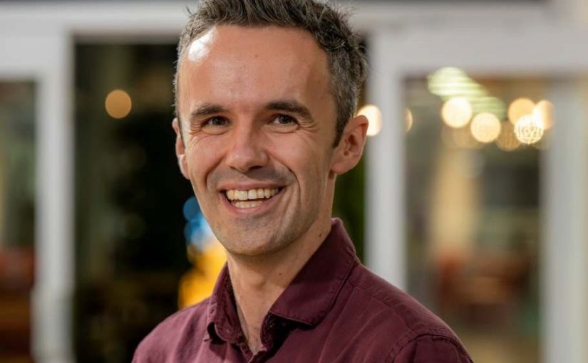 Image of Stuart Cumming. Close-up portrait of a person wearing a burgundy long-sleeve shirt, standing indoors and turned slightly toward the camera. The background is softly blurred with warm, circular lights and glass panels, suggesting a relaxed indoor social or communal environment.
