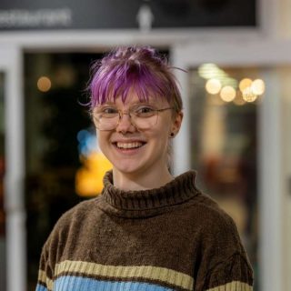 Image of Tilde Resare. Close-up portrait of a person wearing round glasses and a brown striped sweater with blue and beige bands, standing indoors. The background is softly blurred with warm, circular lights and glass panels, indicating a relaxed indoor social setting.