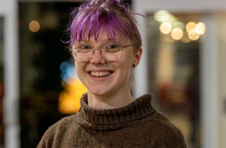 Image of Tilde Resare. Close-up portrait of a person wearing round glasses and a brown striped sweater with blue and beige bands, standing indoors. The background is softly blurred with warm, circular lights and glass panels, indicating a relaxed indoor social setting.