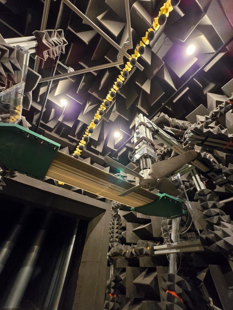 Upward view inside an anechoic chamber lined with dark acoustic foam wedges, showing a mounted propeller on a test rig and a suspended linear array of microphones with yellow foam covers, used for detailed aerodynamic noise measurement.