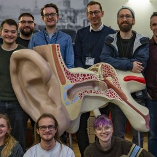 Cohort 1 of the EPSRC Sustainable Sound Futures CDT gathered for a group photo indoors around a large anatomical ear model used for sound and hearing research.
