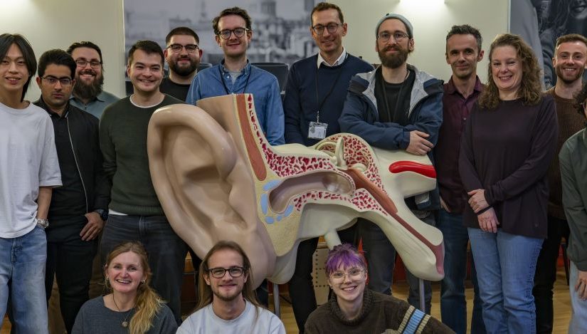 Cohort 1 of the EPSRC Sustainable Sound Futures CDT gathered for a group photo indoors around a large anatomical ear model used for sound and hearing research.