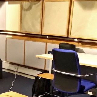 Person seated in an acoustically treated listening room with speakers, monitor, and sound testing equipment.