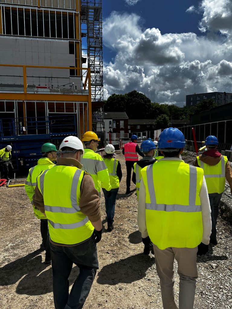 Group of people in PPE and hardhats walking around the building site of at new acoustics building in Salford. The building is partially constructed with scaffolding and external stairwell in the background