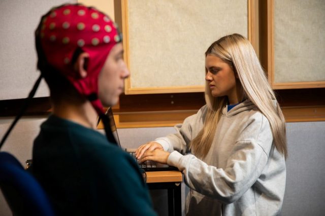 A participant sits wearing a red EEG cap with multiple electrodes while another researcher operates a laptop, inside a laboratory with acoustic panels on the walls for sound perception or neuroscience experiments.