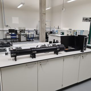 A laboratory workspace with a long cylindrical test apparatus mounted on supports on a bench, alongside a computer, measurement equipment, and a vertical testing rig, set within a clean, well-lit engineering lab.