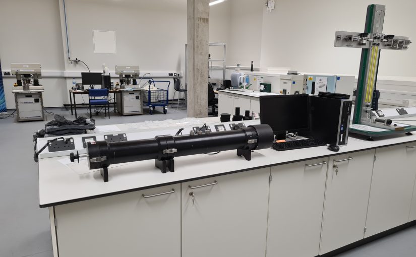 A laboratory workspace with a long cylindrical test apparatus mounted on supports on a bench, alongside a computer, measurement equipment, and a vertical testing rig, set within a clean, well-lit engineering lab.