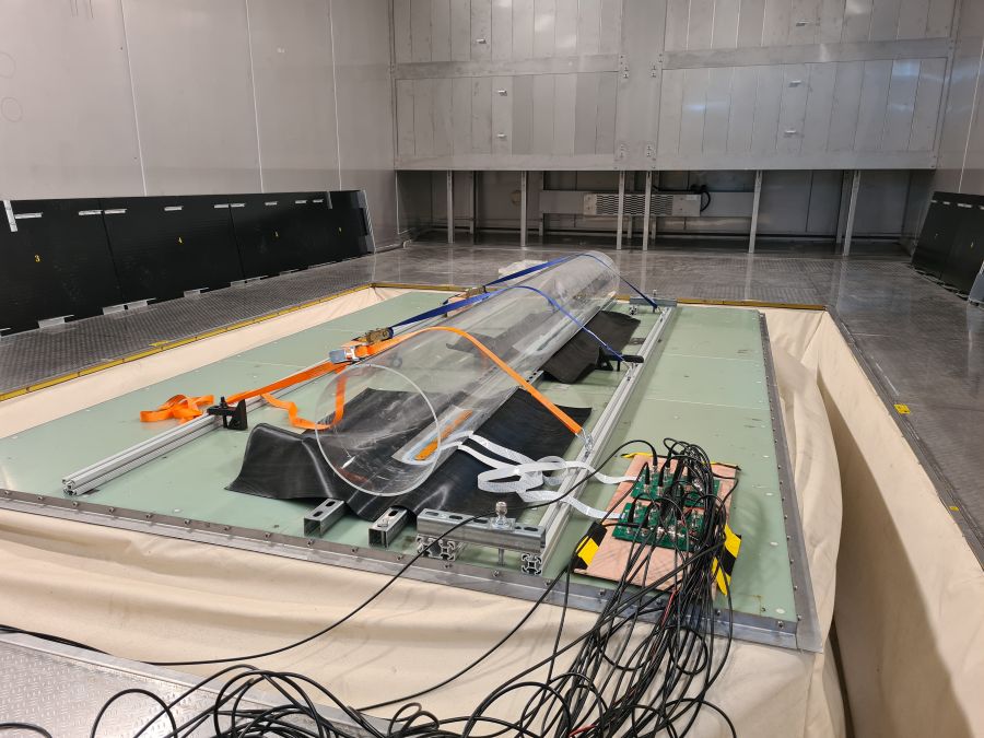 A laboratory wind tunnel test setup featuring a long transparent cylindrical model secured on a platform, with cables, sensors, and data acquisition equipment attached, inside a large industrial testing room with metal flooring and surrounding instrumentation.