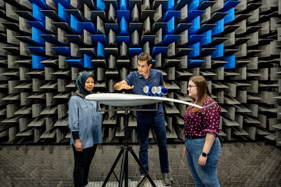 Three researchers stand around an airfoil model mounted on a tripod inside an anechoic chamber lined with blue and grey acoustic foam, as one gestures toward sensors attached to the model during a sound measurement experiment.