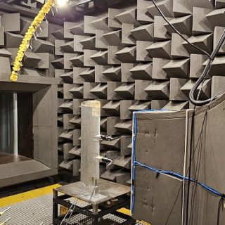 A specialised acoustics testing chamber is lined with large wedge-shaped sound-absorbing foam panels on the walls and ceiling. In the centre of the room, a circular array of microphones mounted on a large ring-shaped frame surrounds experimental equipment positioned on a grated floor. Additional sensors, cables and acoustic measurement devices are connected to a test setup near a large opening in the chamber wall. The facility is designed for precision acoustic measurements in a controlled, reflection-free environment.
